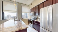 This 2-bedroom, 3-bathroom residence is thoughtfully laid out on a single floor, offering a functional and stylish living environment. The open-concept kitchen and dining area serves as the heart of the home, featuring rich dark-wood cabinetry with gold hardware, stainless steel appliances, a subway tile backsplash, and a generously sized granite island with a built-in sink and pendant lighting overhead. Large windows flood the kitchen with natural light and frame striking urban skyline views. The bedrooms are finished with warm hardwood flooring and roller shades on expansive windows that continue the city view theme. The primary bedroom offers a comfortable footprint with ample closet access. Three closets throughout the unit provide generous storage. The bathrooms are elegantly appointed, with at least one featuring marble-style tile on both the walls and floor, a soaking tub with a glass enclosure, and chrome fixtures. A dedicated laundry room and a connecting hallway complete the layout. Neutral wall tones, white trim, and consistent hardwood flooring throughout create a cohesive, clean aesthetic that is both modern and inviting.