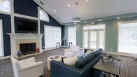 This single-floor communal lounge is a well-appointed gathering space designed with both comfort and style in mind. The room immediately draws attention with its dramatic vaulted ceilings and a striking combination of deep navy and soft teal accent walls, creating a bold yet welcoming atmosphere. An arched transom window crowns the fireplace wall, flooding the space with natural light alongside multiple large windows fitted with crisp white blinds and glass-paneled French doors that open to the outdoors. The fireplace, framed by a classic white mantel, serves as a focal point and adds a warm, residential feel to the room. Seating is thoughtfully arranged around a central coffee table, featuring a tufted navy sofa, white accent chairs, and a round ottoman, all set atop a patterned area rug that anchors the space. A decorative pendant light fixture hangs from the ceiling, complementing the recessed lighting throughout. A high-top bar table with stools is positioned near the windows, offering an additional casual gathering spot. The overall ambiance is polished and inviting, blending classic architectural details with contemporary furnishings to create a versatile and comfortable shared environment.