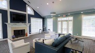 This single-floor communal lounge is a well-appointed gathering space designed with both comfort and style in mind. The room immediately draws attention with its dramatic vaulted ceilings and a striking combination of deep navy and soft teal accent walls, creating a bold yet welcoming atmosphere. An arched transom window crowns the fireplace wall, flooding the space with natural light alongside multiple large windows fitted with crisp white blinds and glass-paneled French doors that open to the outdoors. The fireplace, framed by a classic white mantel, serves as a focal point and adds a warm, residential feel to the room. Seating is thoughtfully arranged around a central coffee table, featuring a tufted navy sofa, white accent chairs, and a round ottoman, all set atop a patterned area rug that anchors the space. A decorative pendant light fixture hangs from the ceiling, complementing the recessed lighting throughout. A high-top bar table with stools is positioned near the windows, offering an additional casual gathering spot. The overall ambiance is polished and inviting, blending classic architectural details with contemporary furnishings to create a versatile and comfortable shared environment.
