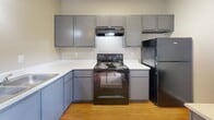 Located at 1623 Aquarena Springs Dr in San Marcos, TX, this one-bedroom, one-bathroom unit offers an efficiently designed single-floor layout. The kitchen is well-appointed with gray cabinetry, white countertops, hardwood-style flooring, and black appliances including an electric range, refrigerator, and dishwasher. A double-basin sink sits beneath a wall-mounted light fixture, and ample upper and lower cabinet storage keeps the space organized and functional. The bedroom features plush carpeting, neutral-toned walls, and a ceiling fan overhead. Large windows invite generous natural light and offer views of the surrounding greenery, while mirrored sliding closet doors add a sense of depth to the room. The bathroom includes a full tub and shower combination with built-in shelving, chrome fixtures, and clean white surround walls. The overall layout is straightforward and practical, making efficient use of the available space. The unit's proximity to Aquarena Springs Drive places it conveniently within the San Marcos community, with mature trees visible from the windows contributing to a pleasant outdoor backdrop.