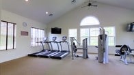 This fitness center offers a bright, welcoming environment designed to support a well-rounded workout routine. The space is defined by its impressive vaulted ceilings, which lend an open, airy quality and create a sense of spaciousness throughout. A ceiling fan mounted at the peak ensures comfortable air circulation during exercise sessions.

Natural light floods the room through a series of windows, including a striking arched window that serves as a focal point on one wall, offering views of the surrounding greenery. The neutral, light-toned walls and carpeted flooring contribute to a clean and comfortable atmosphere.

The cardio area is anchored by a row of three treadmills, each equipped with individual display consoles, positioned facing a wall-mounted flat-screen television — ideal for entertainment during workouts. Alongside the cardio equipment, the space includes a multi-station strength training machine, providing options for a variety of resistance exercises, as well as additional weight and cable training equipment to round out the available fitness tools.

The single-floor layout ensures easy accessibility throughout, and the thoughtful arrangement of equipment allows for ample circulation space between stations. Overall, this fitness center combines functional amenities with an inviting, well-lit environment suitable for residents or members seeking a convenient and comprehensive place to exercise.