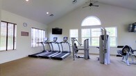 This fitness center offers a bright, welcoming environment designed to support a well-rounded workout routine. The space is defined by its impressive vaulted ceilings, which lend an open, airy quality and create a sense of spaciousness throughout. A ceiling fan mounted at the peak ensures comfortable air circulation during exercise sessions.

Natural light floods the room through a series of windows, including a striking arched window that serves as a focal point on one wall, offering views of the surrounding greenery. The neutral, light-toned walls and carpeted flooring contribute to a clean and comfortable atmosphere.

The cardio area is anchored by a row of three treadmills, each equipped with individual display consoles, positioned facing a wall-mounted flat-screen television — ideal for entertainment during workouts. Alongside the cardio equipment, the space includes a multi-station strength training machine, providing options for a variety of resistance exercises, as well as additional weight and cable training equipment to round out the available fitness tools.

The single-floor layout ensures easy accessibility throughout, and the thoughtful arrangement of equipment allows for ample circulation space between stations. Overall, this fitness center combines functional amenities with an inviting, well-lit environment suitable for residents or members seeking a convenient and comprehensive place to exercise.