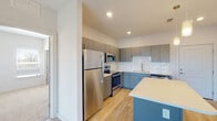 This well-appointed single-floor residence showcases a thoughtfully designed open-concept layout that emphasizes functionality and modern style. The kitchen serves as the heart of the home, featuring two-tone cabinetry — sleek gray upper cabinets paired with blue-accented lower cabinets — all finished with brushed nickel hardware. A full suite of stainless steel appliances, including a refrigerator, range, microwave, and dishwasher, complements the crisp white quartz countertops and classic white subway tile backsplash. A center island with a light countertop and pendant lighting above provides additional prep space and casual seating. Light hardwood-style flooring flows throughout the main living areas, while the kitchen opens naturally into a bright, spacious living area illuminated by windows with exterior views. The bedroom offers plush carpeting, a ceiling fan, and multiple windows that invite natural light. A walk-in closet with wire shelving provides generous storage. The bathroom is finished with white subway tile surrounding the bathtub and shower, wood-look flooring, and a clean white vanity with an undermount sink. The overall aesthetic is contemporary and cohesive, combining neutral tones with modern finishes throughout.