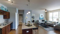 This well-designed single-floor residence showcases a thoughtfully planned open-concept layout that promotes a natural flow between the main living areas. The kitchen serves as a central hub, anchored by a rich dark-wood island topped with granite and illuminated by elegant pendant lights that hang from the ceiling above. The kitchen opens directly into the living area, which features warm hardwood flooring, neutral gray walls, and a ceiling fan that enhances the airy atmosphere. The living space is generously proportioned, offering ample room for comfortable furnishings and day-to-day living. A peek into the bedroom reveals a cozy, well-lit retreat with coordinated décor and a layout that maximizes the available space. Throughout the residence, white panel doors and clean architectural lines provide a polished, cohesive aesthetic. Recessed lighting complements the pendant and ceiling fixtures, ensuring the interior is well-lit throughout. The neutral color palette of soft grays and warm wood tones creates a calm, inviting ambiance, making this a comfortable and functional living space suited for a variety of lifestyles.