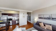 This single-floor unit offers a thoughtfully designed layout that balances comfort and functionality. The open-concept living area is anchored by warm hardwood flooring that extends throughout the space, creating a cohesive and inviting atmosphere. The living room is generously sized, accommodating a full seating arrangement with a sofa, accent chair, and a round coffee table, all set atop a plush area rug. A large window with vertical blinds floods the room with natural light and hints at elevated views beyond. Textured ceilings add a classic architectural detail throughout the unit. The kitchen is efficiently laid out and features rich dark-wood cabinetry, granite countertops, stainless steel appliances including a refrigerator, electric range, and dishwasher, as well as a stainless steel sink. A small dining area adjacent to the kitchen provides a convenient space for everyday meals. The bathroom is clean and well-appointed, featuring a white vanity with under-cabinet storage, a large mirror with overhead lighting, a combination tub and shower with tiled surround, and hardwood flooring that continues from the main living areas. The overall ambiance of the unit is modern and bright, with a neutral color palette of soft grays and white that lends itself to a versatile and welcoming living environment.