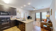 This efficiently designed 1-bedroom, 1-bathroom unit offers a thoughtful single-floor layout that maximizes functionality and comfort. The open-concept kitchen and living area serves as the heart of the space, where rich dark espresso cabinetry contrasts beautifully with light countertops and hardwood-style flooring. The kitchen is well-equipped with a full suite of black appliances, including a refrigerator, range, and microwave, along with ample upper and lower cabinet storage. A peninsula with bar seating extends the counter space and connects the kitchen seamlessly to the living area. The living room receives abundant natural light through multiple windows dressed with floor-length curtains, creating a bright and welcoming atmosphere. A ceiling fan overhead adds to the comfort of the space. The bedroom features plush carpeting, a ceiling fan, and a dedicated closet with built-in shelving, providing practical storage. An additional door from the bedroom leads conveniently to the bathroom, which includes a dark-cabinet vanity with a white countertop, a large mirror, and hardwood-style flooring. A separate closet completes the layout, offering extra storage. The overall design is clean, cohesive, and functional throughout.