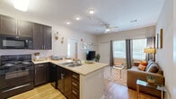 This efficiently designed 1-bedroom, 1-bathroom unit offers a thoughtful single-floor layout that maximizes functionality and comfort. The open-concept kitchen and living area serves as the heart of the space, where rich dark espresso cabinetry contrasts beautifully with light countertops and hardwood-style flooring. The kitchen is well-equipped with a full suite of black appliances, including a refrigerator, range, and microwave, along with ample upper and lower cabinet storage. A peninsula with bar seating extends the counter space and connects the kitchen seamlessly to the living area. The living room receives abundant natural light through multiple windows dressed with floor-length curtains, creating a bright and welcoming atmosphere. A ceiling fan overhead adds to the comfort of the space. The bedroom features plush carpeting, a ceiling fan, and a dedicated closet with built-in shelving, providing practical storage. An additional door from the bedroom leads conveniently to the bathroom, which includes a dark-cabinet vanity with a white countertop, a large mirror, and hardwood-style flooring. A separate closet completes the layout, offering extra storage. The overall design is clean, cohesive, and functional throughout.