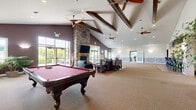 Located at 18 Stoneledge Dr in Troy, NY, this impressive single-floor community clubhouse offers a generously sized, amenity-rich environment designed for recreation and socializing. The centerpiece of the facility is a expansive great room with soaring vaulted ceilings accented by exposed wooden beam details and multiple ceiling fans. A striking floor-to-ceiling stacked stone fireplace anchors one end of the room, complemented by large windows that flood the space with natural light and offer views of the surrounding greenery. Comfortable seating areas invite relaxation, while plush carpeting adds warmth throughout. Adjacent to the great room is a game area featuring a full-size billiards table, and a separate room houses additional entertainment options including an air hockey table, arcade games, and a dartboard with easy-care hardwood flooring. A well-appointed kitchen with dark cabinetry, stainless steel appliances, and a bar with counter seating provides an ideal space for gatherings. The dedicated exercise room is fully equipped with treadmills, ellipticals, a stationary bike, free weights, and a weight bench, all illuminated by large windows overlooking lush trees and complemented by full-wall mirrors and mounted televisions.