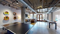 This single-floor amenity space showcases a bold industrial design aesthetic, characterized by exposed concrete ceilings, visible ductwork and piping, polished concrete floors, and painted concrete block walls. The open layout seamlessly connects multiple functional zones, creating a versatile environment suited for both recreation and relaxed socializing.

The game area anchors one end of the space with a full-size ping pong table and additional game furnishings, set beneath dramatic exposed mechanical systems. Adjacent to it, a long natural wood communal dining table with wood-and-metal chairs sits beside a sleek kitchenette outfitted with deep navy cabinetry and a sink — ideal for casual dining or group gatherings.

The lounge area features generously sized sectional sofas arranged around low coffee tables, accented by vibrant, circular repurposed street sign wall art that adds a playful, urban character. A wall-mounted flat-screen television completes the entertainment setup, while colorful multi-toned carpet tiles define and soften the seating zone.

Large floor-to-ceiling glass doors and expansive windows flood the interior with natural light and open directly to an outdoor patio. The exterior area features paved stone flooring, seating arrangements, and colorful outdoor furniture, offering a comfortable extension of the interior. Pendant lighting and a gated entry complete the outdoor space, which is framed by the building's striking brick and concrete façade.