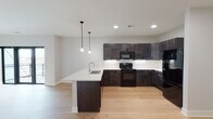 This single-floor residence showcases a contemporary design with a cohesive color palette and quality finishes throughout. The kitchen serves as a focal point, featuring rich dark-stained cabinetry extending to the upper walls, a classic white subway tile backsplash, and a full suite of black appliances including a range, over-the-range microwave, and refrigerator. A peninsula with a light quartz countertop and an undermount sink provides additional workspace and casual dining potential, accented by two sleek pendant lights overhead. Light-toned hardwood flooring flows through the kitchen and adjoining living area, creating a warm, cohesive aesthetic.

The open layout connects the kitchen to a generously sized living area, with a window allowing natural light and views of the surrounding neighborhood. The bedrooms are carpeted in neutral tones and each features a large window with exterior views, providing brightness and a comfortable atmosphere.

Two bathrooms serve the unit, each finished with light tile flooring and dark vanity cabinetry paired with white countertops. One bathroom includes a walk-in shower, while the second offers a full tub and shower combination, both appointed with matte black hardware accents that complement the overall modern aesthetic.