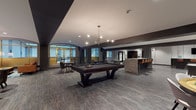 Located on Euclid Avenue in the heart of Cleveland, this amenity floor seamlessly blends historic architecture with contemporary interior design. The centerpiece of the space is a generous open-plan lounge and game room, anchored by a full-size pool table beneath a striking brass multi-arm pendant light. Wood-look flooring and dark exposed beams contrast with light walls to create a sophisticated, urban aesthetic throughout.

Adjacent to the game area, a dedicated lounge nook features a pair of cognac leather sofas flanking a chevron-patterned wood coffee table, with a wall-mounted fireplace and built-in shelving serving as a stylish focal point. A wall-mounted television completes the entertainment setup.

The kitchenette area is well-appointed with granite countertops, muted sage cabinetry, and a coffee station, providing a functional refreshment hub. Arched gold-framed windows offer views into the outdoor courtyard, adding architectural character and natural light.

The expansive courtyard, enclosed by the building's classic red brick facade with copper-trimmed detailing, offers generous outdoor seating with multiple lounge and dining arrangements on a tiled terrace. A wide corridor connects the interior zones, finished with recessed lighting and dark-stained doors with gold hardware, maintaining the space's cohesive, modern-classic design language.