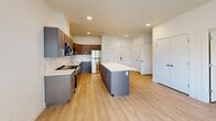 Located at 105 S Main St in Layton, Utah, this single-floor unit offers a thoughtfully designed open-concept layout that maximizes its compact footprint. The kitchen serves as the centerpiece of the living area, showcasing a large central island with a crisp white countertop, sleek gray cabinetry, and a full suite of stainless steel appliances including a gas range, microwave, and refrigerator. A white subway tile backsplash adds a clean, modern touch, while three pendant lights hang above the island, providing both style and task lighting. Light wood-look flooring runs throughout the entire unit, lending warmth and visual continuity. The main living area is bright and airy, benefiting from multiple windows and a glass-panel exterior door that invite abundant natural light. The bathroom is well-appointed with a dual-sink vanity featuring gray cabinetry and a large mirror, and conveniently incorporates a stacked washer and dryer unit. Two closets offer practical storage solutions, including a walk-in closet with wrap-around shelving and hanging rods. Clean white walls and recessed lighting throughout contribute to the unit's fresh, contemporary aesthetic, making it a well-equipped and efficient living space.