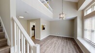 Located at 1009 Fir Pl in Greensboro, NC, this two-story home showcases a thoughtfully designed interior with a neutral color palette and light wood-look flooring carried throughout both levels. The main living area immediately impresses with its dramatic double-height ceiling, which creates an expansive, airy atmosphere. A contemporary metal chandelier serves as a central focal point, complementing the open layout below. A white-railed staircase connects the two floors, while an upper-level overlook adds architectural depth to the space.

Large windows with deep sills flood the living area with natural light and offer views of the surrounding greenery, enhancing the open feel. A pass-through window connects the living area to the kitchen, maintaining visual connectivity between spaces. The galley-style kitchen is well-appointed with white cabinetry, quartz-style countertops, and stainless steel appliances, including a range, microwave, refrigerator, and dishwasher.

The upper level features a barn door accent that adds a modern farmhouse touch, alongside an open landing overlooking the main floor. Bathrooms include a full tub-shower combination and a secondary bath with a floating wood-toned vanity and angled ceiling detail. The overall ambiance is clean, bright, and move-in ready.