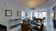 This single-floor space showcases a thoughtfully designed open-plan layout that seamlessly connects the dining area to the living space and bedroom beyond. The dining area serves as a central focal point, anchored by a glass-top dining table surrounded by rich cognac leather chairs, creating an inviting yet sophisticated atmosphere. Overhead, a dramatic multi-armed brass chandelier with exposed bulbs adds a contemporary artistic element to the space.

The light-toned walls and pale flooring contribute to a bright, airy ambiance, further enhanced by floor-to-ceiling windows that frame impressive urban skyline views, flooding the interior with natural light. A sleek console table with metal X-frame detailing lines one wall, styled with decorative accessories and framed artwork that lend a curated, gallery-like quality to the space.

Through open doorways, a glimpse of the bedroom reveals a neatly appointed sleeping area with an upholstered headboard, maintaining the same refined aesthetic throughout. The living area transitions smoothly from the dining space, reinforcing the open, flowing nature of the floor plan. Overall, the space balances modern design with warm tones for an elegant, urban feel.