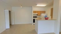 This single-floor space offers a clean, open-concept layout that seamlessly connects the kitchen and living area, creating a comfortable and functional environment. The living area is bright and airy, featuring light-toned tile flooring that extends throughout the space, contributing to a cohesive and easy-to-maintain interior. White walls and a neutral color palette give the space a fresh, welcoming feel, while a track light fixture provides focused overhead lighting in the living zone.

The kitchen is efficiently designed and positioned adjacent to the living area, separated by a peninsula-style countertop that doubles as a casual breakfast bar. Warm-toned wood cabinetry lines the upper walls, offering ample storage space. The kitchen is equipped with standard appliances, including a white electric range with an overhead exhaust hood, a refrigerator, and a sink integrated into the countertop. A flush-mounted fluorescent light fixture illuminates the kitchen workspace, ensuring practical visibility for cooking and meal preparation.

The open flow between the kitchen and living space makes this layout particularly well-suited for everyday living and casual entertaining. The overall design is straightforward and functional, with a simple, understated aesthetic that provides a versatile canvas for personalization.