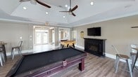 This single-floor game room is designed for entertainment and social gatherings, combining generous open space with a well-appointed layout. The standout architectural feature is the vaulted tray ceiling, which adds a sense of height and airiness to the room. Dual ceiling fans with recessed lighting ensure comfort and visibility throughout the space.

The main area centers around a full-sized pool table with a dark felt surface and richly stained wood frame, complemented by a foosball table nearby. A wall-mounted flat-screen television sits above a sleek black electric fireplace, creating a natural focal point for relaxed viewing. Industrial-style metal bar stools and wooden pub tables provide casual seating options throughout the room.

Natural light floods the space through multiple sets of French doors along one wall, offering views of the surrounding outdoor greenery. A smaller seating nook near a transom-topped door provides a quieter corner for conversation.

A kitchenette, finished with dark cabinetry, a subway tile backsplash, and stainless steel appliances including a refrigerator and range, supports food and beverage preparation for gatherings. Warm hardwood-style flooring and neutral taupe walls tie the entire space together with a clean, welcoming aesthetic.