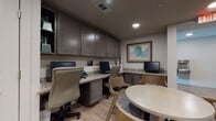 This single-floor commercial common area is thoughtfully designed to serve as both a functional business center and a welcoming lounge space. The business center features multiple built-in workstations set along granite-topped counters, complemented by dark espresso cabinetry that extends to the ceiling for ample storage. Ergonomic task chairs and desktop computers at each station make the space fully equipped for productivity. A rounded table with modern curved chairs provides an additional collaborative seating area, ideal for informal meetings or group work.

The flooring throughout is a warm wood-look tile that adds a cohesive, contemporary feel to the space. Recessed lighting illuminates each area effectively, while decorative artwork — including a vibrant teal abstract canvas and geometric wood-panel wall art — adds visual interest and a polished aesthetic. A circular sculptural wall accent and a bold large-format abstract painting near a cozy lounge chair with an accent lamp further elevate the space's design character.

Accessible restrooms are conveniently located off the main corridor, clearly marked and ADA-compliant. A water fountain is also visible in the connecting hallway. The overall ambiance is clean, modern, and professional, balancing utility with thoughtful interior design details.