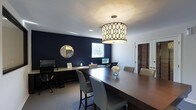 This single-floor space presents a polished and professional interior centered around a well-appointed meeting or conference room. The room makes an immediate visual impact with its deep navy accent wall, which serves as a striking backdrop against the crisp white ceiling and trim. A large, dark wood conference table dominates the space, surrounded by sleek, upholstered chairs in a neutral grey tone, offering both comfort and a refined aesthetic.

Overhead, a statement drum pendant light with a geometric patterned shade provides warm, focused illumination, complementing the recessed lighting integrated into the white ceiling. A decorative sunburst mirror adorns the navy wall, adding a touch of elegance to the space. Along one side of the room, a built-in or freestanding desk area is equipped with computer monitors, suggesting a functional workspace that supports both meetings and individual work tasks.

Natural light filters in through a window on the navy wall, as well as through a shuttered window near the entrance door, adding brightness and balance to the room. A small floral arrangement at the center of the table adds a welcoming, finishing touch to this smart and sophisticated space.