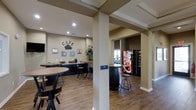 This café is a thoughtfully designed single-floor gathering space that combines functionality with a warm, inviting atmosphere. The interior features warm beige walls accented by decorative wall art and a charming metal "CAFE" sign, establishing a casual yet polished ambiance throughout. Hardwood-style flooring runs across the entire space, complementing the dark cabinetry and furniture for a cohesive aesthetic.

The seating area is furnished with high-top round tables and adjustable bar-height stools, offering a relaxed environment well-suited for socializing or a quiet break. Structural columns with crown molding detailing add architectural character while defining the open layout. Recessed lighting illuminates the space evenly, enhancing the airy feel of the room.

Along one wall, a dedicated refreshment counter houses a single-serve coffee maker and accompanying supplies, providing a convenient self-serve beverage station. Adjacent to this area, a central barrel-style high-top table with bar stools creates an additional focal seating point. A vending machine station, visible through a connecting passage, offers snack and beverage options. A glimpse of fitness equipment in the background suggests this café is part of a larger amenity facility, making it an ideal hub for residents or visitors seeking refreshment and community connection.