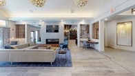 Located at 100 Gateway in Scarborough, Maine, this single-floor amenity space is thoughtfully designed to foster comfort, community, and functionality. The open-concept layout flows seamlessly between distinct zones, each offering its own character while contributing to a cohesive, modern aesthetic.

The central lounge area serves as the heart of the space, featuring a sleek linear fireplace flanked by stone tile surround, a wall-mounted television, and an inviting arrangement of contemporary sofas and accent chairs set atop patterned area rugs. Light wood-look tile flooring runs throughout, lending warmth and continuity. Decorative globe pendant lights and recessed lighting illuminate the space with a layered glow.

A standout lounge nook showcases a rich wood slatted ceiling, a curved sectional sofa in a teal hue, and an additional fireplace — creating a cozy, intimate retreat within the larger communal setting. Wooden slat partition walls add architectural texture and define zones without fully enclosing them.

The café and bar area features granite countertops, warm wood cabinetry, a curved island, and a wood-paneled ceiling with pendant lighting, equipped for beverage service. Adjacent meeting and dining rooms offer round and rectangular tables suited for small gatherings or collaborative work, with large windows that draw in natural light. Curated artwork adorns the walls, adding a polished, residential feel throughout.