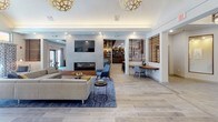 Located at 100 Gateway in Scarborough, Maine, this single-floor amenity space is thoughtfully designed to foster comfort, community, and functionality. The open-concept layout flows seamlessly between distinct zones, each offering its own character while contributing to a cohesive, modern aesthetic.

The central lounge area serves as the heart of the space, featuring a sleek linear fireplace flanked by stone tile surround, a wall-mounted television, and an inviting arrangement of contemporary sofas and accent chairs set atop patterned area rugs. Light wood-look tile flooring runs throughout, lending warmth and continuity. Decorative globe pendant lights and recessed lighting illuminate the space with a layered glow.

A standout lounge nook showcases a rich wood slatted ceiling, a curved sectional sofa in a teal hue, and an additional fireplace — creating a cozy, intimate retreat within the larger communal setting. Wooden slat partition walls add architectural texture and define zones without fully enclosing them.

The café and bar area features granite countertops, warm wood cabinetry, a curved island, and a wood-paneled ceiling with pendant lighting, equipped for beverage service. Adjacent meeting and dining rooms offer round and rectangular tables suited for small gatherings or collaborative work, with large windows that draw in natural light. Curated artwork adorns the walls, adding a polished, residential feel throughout.