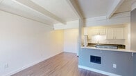 Located at 1200 West Gonzales Road in Oxnard, California, this single-floor one-bedroom unit offers a thoughtfully arranged layout across a compact but functional footprint. The interior is unified by warm wood-tone laminate flooring and crisp white walls throughout, creating a clean, bright aesthetic.

The living area is open and uncluttered, benefiting from the architectural detail of exposed ceiling beams that add character to the otherwise modern, minimalist design. A pass-through counter connects the living space to the kitchen, enhancing the sense of openness while maintaining a defined cooking zone.

The kitchen is efficiently designed in a galley configuration, featuring white cabinetry with brushed hardware, dark granite-style countertops, a white appliance suite including a range and refrigerator, and ample upper and lower storage. Under-cabinet lighting adds a practical and polished touch.

The primary bedroom is generously proportioned relative to the overall unit, offering room for a full furniture arrangement. It features a mirrored sliding closet, a window with vertical blinds that admits natural light, and the same warm flooring found throughout the space. Exposed ceiling beams carry into this room, maintaining design continuity.

The layout flows intuitively from the living and kitchen areas into the bedroom, with clean transitions and consistent finishes making the space feel cohesive and well-maintained.