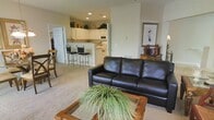 This single-floor residence is designed with an open-concept layout that seamlessly connects the living and kitchen areas, creating a spacious and cohesive atmosphere. The living room serves as the central gathering space, featuring neutral-toned carpeting and recessed lighting that lend a clean, understated aesthetic. A dark leather sofa anchors the seating area alongside a wood-framed coffee table, while decorative accents — including framed wall art and a wooden cabinet with display pieces — add character to the space.

The kitchen is positioned toward the rear and is outfitted with white cabinetry, white appliances, and a raised breakfast bar with two bar stools, making it practical for casual dining or conversation while cooking. Greenery atop the upper cabinets adds a touch of warmth to the crisp, neutral palette. A doorway near the kitchen leads to additional rooms, suggesting a functional flow throughout the residence. Recessed lighting in both areas ensures consistent, even illumination. Overall, the space balances practicality with a comfortable, welcoming ambiance suitable for everyday living.