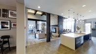 This single-floor space masterfully blends rustic charm with modern sophistication through a thoughtfully curated open-concept layout. Exposed dark wood beams run across the ceiling, adding warmth and architectural character, while bold structural columns clad in dark metal anchor the space with an industrial edge. A floor-to-ceiling stone fireplace serves as a dramatic focal point, framed by dark accent walls that create a striking contrast against the lighter surroundings. The living area features light hardwood flooring and is furnished with a neutral-toned seating arrangement, creating a comfortable and inviting atmosphere. Recessed lighting throughout ensures a well-lit environment, complemented by the ample natural light streaming in through large windows that offer views of the outdoors. The adjacent area transitions to large-format tile flooring, suggesting a functional zone such as a kitchen or reception space, with pendant lighting adding a refined touch overhead. The overall ambiance is warm yet polished, combining natural materials like stone and wood with sleek finishes to achieve a balanced, contemporary aesthetic. This space is well-suited for both relaxed gatherings and more formal settings.