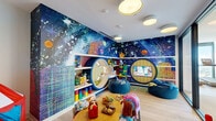 Located at 700 Northeast 1st Avenue in Miami, FL, this imaginative children's playroom delivers a fully immersive environment designed to spark creativity and play. The room's most defining feature is a sweeping, floor-to-ceiling galaxy mural that wraps around all four walls, depicting vibrant planets, stars, and cosmic scenery in bold blues, oranges, and greens, instantly transporting young visitors into an outer space adventure.

Built-in white shelving lines one wall, offering organized storage for colorful building blocks, stuffed animals, and books. Integrated into the shelving are two circular porthole-style enclosures framed with chevron-patterned fabric — one functioning as a cozy reading nook and the other housing a wall-mounted display screen for entertainment. Plush bean bag chairs and a city-map play rug provide comfortable, casual seating areas throughout.

A child-sized round table with chairs serves as a central activity area, complemented by a whimsical pop-up circus tent that adds a playful focal point. Light wood flooring runs throughout, contributing warmth and easy maintenance, while flush-mounted ceiling lights ensure bright, even illumination. A full-height glass partition wall offers a transparent view directly into an adjacent, well-equipped fitness center, connecting the two amenity spaces while maintaining acoustic separation.