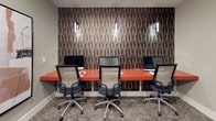 This single-floor business center is thoughtfully designed to support focused work in a polished, contemporary environment. The space is immediately distinguished by its curated selection of large-scale abstract artwork, displayed prominently throughout the corridors and work areas. These bold pieces — featuring expressive brushstrokes in warm terracotta, black, and neutral tones — add a gallery-like quality that elevates the overall ambiance.

The main work area features a continuous orange worksurface that seats multiple users side by side, each station equipped with a desktop computer. Pendant lighting drops down above the workstations against a richly textured, geometric feature wall in deep browns and golds, creating a striking visual backdrop while maintaining an atmosphere conducive to concentration.

A dedicated printing and support station is positioned nearby, complete with a printer and shelving, offering functional convenience within easy reach. The space also includes a private room accessed through a bold navy-blue door, which provides an option for more secluded work or meetings.

Throughout the space, patterned carpet in muted tones grounds the design, while recessed lighting ensures consistent, even illumination. Navy accent doors with paneled detailing appear consistently across the space, tying the design language together with a cohesive and refined aesthetic.