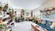 This single-floor unit offers a warm and eclectic living experience defined by abundant greenery, colorful décor, and a thoughtfully lived-in character. The living area serves as the heart of the space, featuring plush carpeted flooring, a textured ceiling, and multiple windows that invite natural light from multiple directions. A wall-mounted air conditioning unit provides climate control, while a baseboard heating system runs along the perimeter. The room accommodates distinct seating zones, including a papasan chair nestled in a sunlit reading corner surrounded by an impressive collection of potted plants, and a teal futon sofa accented with vibrant throw pillows beneath a bold canvas artwork. A mid-century modern coffee table anchors the seating area, complementing the eclectic aesthetic. A dedicated dining nook with a small table and chairs is also present within the living space. The compact kitchen features light-toned cabinetry, laminate countertops, a double-basin sink, and a freestanding electric range, with a window above the counter adding natural light. The bedroom is an intimate retreat adorned with an ornate mandala tapestry that serves as a dramatic focal point above the bed. The bathroom includes tile-clad walls, stone-look flooring, a pedestal sink, a framed mirror, and a window sill lined with small plants.