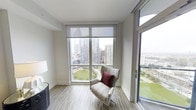 This contemporary high-rise residence offers a refined living experience defined by clean lines, modern finishes, and an abundance of natural light. The single-floor layout is thoughtfully designed to maximize both comfort and function.

The living area is a standout feature, showcasing expansive floor-to-ceiling windows that frame breathtaking views of the city skyline and a lush green park below. Large sliding glass doors lead to an outdoor balcony, further blurring the line between indoor and outdoor living. Light wood-tone flooring runs throughout, complementing the neutral white walls and creating a warm, airy atmosphere. Roller shades offer flexible light control while preserving the dramatic vistas.

The space is styled with contemporary furnishings that reflect its modern character — illustrated by a patterned accent chair positioned to take full advantage of the panoramic outlook. The overall ambiance is one of understated sophistication, with a palette of soft neutrals accented by rich tones that add depth and warmth. Whether enjoying the cityscape by day or the glittering skyline by night, this residence offers an elevated urban living experience defined by light, views, and modern design.