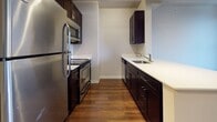 This well-appointed single-floor unit offers a cohesive modern design throughout, anchored by rich, dark hardwood flooring that extends seamlessly from the entry corridor through the main living areas. The galley-style kitchen is a functional standout, featuring dark espresso cabinetry paired with crisp white countertops and a full suite of stainless steel appliances, including a refrigerator, range, microwave, and sink with a contemporary faucet. Exposed ductwork along the ceiling adds an industrial loft character, complemented by track lighting that provides focused illumination throughout the kitchen and hallway. The corridor leading from the entry is lined with multiple closet doors, offering generous built-in storage. The bathroom is finished with light stone-look tile flooring, dark vanity cabinetry with a white countertop, and a glass-enclosed walk-in shower, creating a clean and practical space. The bedroom benefits from a large, dark-framed window that fills the room with natural light and frames views of the surrounding urban architecture. The open layout doll's-eye view reveals a compact yet efficiently organized floor plan, making excellent use of the available space with clearly defined kitchen, living, and sleeping zones.