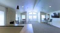 This single-floor space offers an open and airy atmosphere defined by its striking vaulted ceilings that rise to a dramatic peak, creating a strong sense of volume and openness. A large arched window serves as a focal point at the far end of the main room, framing outdoor views and drawing abundant natural light deep into the interior. Additional windows along the surrounding walls further brighten the space throughout the day.

The main gathering area features neutral carpeted flooring and is furnished with casual high-top and standard tables with chairs, making it well-suited for both relaxed lounging and informal meetings. A prominent stone fireplace anchors one wall, adding a warm, textural element to the otherwise light and clean interior. Recessed lighting in the ceiling complements the natural light and ensures the space remains well-lit at any hour.

A countertop surface visible in the foreground suggests a service or refreshment area near the entrance, adding a functional convenience to the space. The overall design palette is understated and neutral, with light-toned walls and trim that enhance the sense of brightness. The combination of architectural interest, natural light, and comfortable furnishings makes this a welcoming and versatile communal environment.