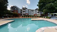 Located at 8810 Autumn Winds Dr in Raleigh, NC, this single-level community amenity space is designed to offer residents a welcoming outdoor retreat with a variety of social and recreational features. The centerpiece of the space is a generously sized resort-style swimming pool with sparkling turquoise water, surrounded by a broad pool deck lined with lounge chairs and shaded by umbrellas, ideal for relaxing on warm Carolina days. The multi-story residential buildings with classic gray siding, brick accents, and balconies frame the pool area, creating an enclosed, community-oriented atmosphere. A covered porch with a herringbone-pattern brick tile floor extends from the clubhouse, supported by crisp white columns and accented with wall-mounted lantern-style lighting. This porch area provides comfortable outdoor seating, including rattan furniture with vibrant green cushions and colorful throw pillows, set against large multi-pane windows that lend a bright, open feel. Adjacent to the covered porch, an open-air concrete patio offers additional seating with high-top tables and chairs, surrounded by blooming crape myrtle trees and lush greenery. Bold decorative planters add pops of color throughout the outdoor spaces. The overall ambiance is lively yet relaxed, blending traditional architectural details with thoughtfully arranged outdoor living areas.