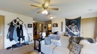 This single-floor residence centers around an open-concept living area that flows naturally toward the kitchen and dining spaces. The living room is carpeted and outfitted with a ceiling fan that provides both lighting and ventilation. A plush, light-colored sofa anchors the seating area and is accompanied by a black side table holding a small lamp and fresh floral arrangement, adding a welcoming touch to the space. The walls display an eclectic mix of artwork, including a large decorative tapestry, contributing to the room's personalized character. A dedicated entryway area near the front features wall hooks and storage for outerwear and accessories. Through the open passageway, the dining area is visible with a dark wood table and chairs, and beyond that, the kitchen is accessible with dark appliances. Additional doorways hint at further rooms, including what appears to be a bathroom with visible lighting. The layout offers a practical and connected flow between the living, dining, and kitchen areas, making the space functional for everyday living on a single level.