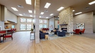 Located at 17309 40th Avenue NE in Arlington, Washington, this single-floor community facility offers a thoughtfully designed layout with a variety of functional and social spaces. The grand entrance lobby features soaring double-height ceilings, large clerestory windows, polished large-format tile flooring, and a built-in reception counter, creating an impressive and welcoming first impression. Adjacent to the lobby, an expansive open lounge area showcases light hardwood flooring, generous skylights, and a dramatic floor-to-ceiling stacked-stone fireplace that serves as a central focal point. This area transitions into a casual dining nook with a wood table, red upholstered chairs, and tall windows offering views of the surrounding residential courtyard. A full kitchen with dark cabinetry, a gas range, white countertops, and a breakfast bar with red barstools supports catering and community dining needs. Additional lounge areas feature plush seating, wall-mounted televisions, and curated artwork throughout. A carpeted game room includes a pool table and accent seating, while a quieter sitting alcove offers comfortable yellow lounge chairs and framed artwork, ideal for relaxed retreat. Natural light permeates the entire space through skylights and large windows, contributing to a bright, welcoming community atmosphere throughout.