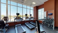 This fitness center is designed to provide a functional and energizing workout environment. The space is defined by a striking wall of floor-to-ceiling windows along one side, flooding the interior with generous natural light and offering open views of the surrounding outdoor area, creating an airy and motivating atmosphere during exercise.

The room features a dark rubberized flooring surface well-suited for gym use, providing both durability and traction. Bold terracotta-red accent walls add a dynamic visual contrast to the neutral tones of the drop-tile ceiling, which houses recessed fluorescent lighting to supplement natural light.

Several treadmills are positioned directly in front of the large windows, each equipped with individual monitor screens for entertainment during cardio workouts. A dedicated free weights area is situated along the adjacent wall, outfitted with a rack of dumbbells in varying sizes to accommodate different training needs. A mirrored wall panel near the weights section enhances the sense of space and supports proper form during strength training. Overall, the fitness center is thoughtfully laid out to support a range of exercise routines in a bright, well-appointed setting.