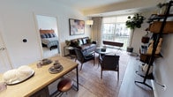This thoughtfully designed single-floor residence blends contemporary style with functional living across a well-organized floor plan. The open-concept living and dining area features light wood-tone flooring, a large sliding glass door that floods the space with natural light, and direct access to a private balcony overlooking the surrounding community. The living area is anchored by a rich leather sofa, a round marble-top coffee table, and a bold abstract artwork accented by a stylish articulating wall sconce.

The kitchen is outfitted with full-height dark wood-grain cabinetry, white quartz countertops, a subway tile backsplash, and a complete suite of stainless steel appliances including a gas range, built-in microwave, refrigerator, and dishwasher. A central island with a dark countertop provides additional prep and dining space.

The primary bedroom offers a serene retreat with a striking four-poster bed frame, soft carpeting, large windows with exterior views, and access to a spacious en-suite bathroom featuring a dual-sink vanity with dark cabinetry, a glass-enclosed walk-in shower, and light tile flooring. A generously sized walk-in closet with adjustable shelving and chrome rods provides ample storage. The secondary bedroom is equally comfortable, served by a full bathroom with a tub-shower combination and clean white finishes throughout.