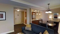 This thoughtfully designed single-floor suite offers a seamless blend of comfort and functionality. The open-concept living area is anchored by a plush navy blue sofa accented with decorative throw pillows, creating an inviting space to relax. Crown molding trims the ceilings throughout, adding a refined, classic touch to the overall aesthetic.

The kitchen is fully equipped and features warm wood cabinetry, granite-style countertops, and stainless steel appliances, including a full-size refrigerator. A breakfast bar with pendant lighting overhead provides a casual dining option, with seating for multiple guests. The pendant lights, finished in a traditional style, contribute to the suite's warm and welcoming ambiance.

Adjacent to the kitchen, a compact dining area offers additional seating for meals. The entryway is clean and functional, with a flush-mount ceiling light and a keycard-access door, indicative of a professional hospitality setting. Wall-unit climate control is conveniently located near the entrance for personalized comfort.

Dark hardwood-style flooring runs throughout the space, grounding the warm neutral wall tones and creating a cohesive, polished look. Overall, this suite offers a comfortable and practical layout suited for extended stays or relaxed, hotel-style living.