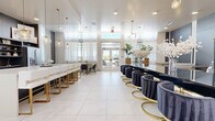 This upscale beauty salon is designed with a refined, contemporary aesthetic that seamlessly blends glamour with functionality. The open-plan layout is anchored by large-format marble-style porcelain tile flooring that flows throughout the entire space, creating a cohesive and polished foundation.

The main service area features two prominent nail bar counters — one finished in sleek black and one in crisp white — each lined with elegantly appointed seating. The black counter is paired with deep navy velvet barrel chairs set on brushed gold bases, while the white counter is accompanied by white upholstered stools with gold-toned frames. Statement globe pendant chandeliers and sputnik-style light fixtures hang overhead, casting a warm, inviting glow throughout.

Along one wall, individual service stations are set against richly textured dark wood paneling, each with its own mirror, task lighting, and plush white armchair seating — offering a more intimate service experience. Tall indoor palms and oversized arrangements of dried white blooms add organic softness and contrast to the crisp interior palette.

A dedicated kitchenette, outfitted with dark wood cabinetry, open shelving, and essential appliances, is neatly integrated into the layout. At the entrance, floor-to-ceiling glass doors flood the reception area with natural light, complemented by sheer white drapery that lends an airy, boutique-hotel ambiance to the entire space.