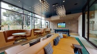 Located at 480 San Antonio Rd in Mountain View, California, this single-floor clubroom is a thoughtfully designed social and recreational space. Floor-to-ceiling glass walls flood the interior with natural light while offering unobstructed views of an outdoor pool courtyard lined with palm trees and yellow umbrellas, creating a seamless indoor-outdoor connection.

The lounge area is anchored by a generous amber-toned sectional sofa adorned with patterned accent pillows, positioned beneath striking geometric chandelier installations that cascade from a deep-teal ceiling. Curved, channel-tufted leather banquette booths line the window wall, each paired with round gold-trimmed tables and sleek gold dome accent lamps, providing intimate seating nooks with garden views.

A dedicated games zone features a full-size billiards table on light hardwood flooring, framed by acoustically textured wood-panel walls and ceiling tiles that add warmth and dimension. A large wall-mounted display screen serves both the games and lounge areas.

A compact kitchenette, finished in dark cabinetry with a clean white countertop, is tucked into the textured wood-paneled corner and equipped with a microwave, beverage tap, and direct access to the outdoor terrace. The overall ambiance blends contemporary design with inviting comfort, making this space ideal for socializing and recreation.