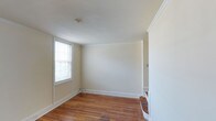 This multi-story property spans three floors, offering a well-structured layout with a classic and understated aesthetic throughout. The interior showcases warm hardwood flooring that adds character and continuity across the living spaces, complemented by crisp white baseboards and elegant crown molding that frame each room with a refined architectural detail.

The rooms are finished in soft, neutral tones that create a bright and airy atmosphere, enhancing the sense of openness throughout the home. Natural light filters in through double-hung windows fitted with blinds, contributing to the welcoming ambiance of each space. The clean, simple wall surfaces provide a versatile backdrop, ready to accommodate a wide range of furnishing styles and personal touches.

The staircase, visible from the main living area, features classic wooden steps that connect the three levels seamlessly, reinforcing the home's traditional character. The layout across multiple floors allows for a natural separation of living, sleeping, and utility spaces. Overall, the property presents a well-maintained, classically styled environment with enduring architectural details that speak to both comfort and functionality.
