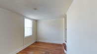 This multi-story property spans three floors, offering a well-structured layout with a classic and understated aesthetic throughout. The interior showcases warm hardwood flooring that adds character and continuity across the living spaces, complemented by crisp white baseboards and elegant crown molding that frame each room with a refined architectural detail.

The rooms are finished in soft, neutral tones that create a bright and airy atmosphere, enhancing the sense of openness throughout the home. Natural light filters in through double-hung windows fitted with blinds, contributing to the welcoming ambiance of each space. The clean, simple wall surfaces provide a versatile backdrop, ready to accommodate a wide range of furnishing styles and personal touches.

The staircase, visible from the main living area, features classic wooden steps that connect the three levels seamlessly, reinforcing the home's traditional character. The layout across multiple floors allows for a natural separation of living, sleeping, and utility spaces. Overall, the property presents a well-maintained, classically styled environment with enduring architectural details that speak to both comfort and functionality.