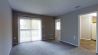 This single-floor residence offers a clean, functional layout designed for comfortable everyday living. The main living area is a standout feature, showcasing wall-to-wall carpeting in a soft neutral tone that adds warmth underfoot. Large sliding glass doors dominate one wall, fitted with vertical blinds that allow for adjustable natural light and direct access to an outdoor area. The abundance of natural light filtering through the sliding doors creates an open, airy atmosphere throughout the room. The walls are finished in a contemporary gray tone, complementing the overall neutral color palette of the space. A floor-mounted HVAC vent ensures year-round climate comfort. White baseboards provide a crisp contrast against the gray walls, adding a subtle architectural detail to the room. A doorway leads to an adjacent room, also finished in matching neutral tones, suggesting a cohesive design language carried throughout the home. The ceiling is smooth and painted white, contributing to the bright and open feel of the interior. Overall, this space presents a well-maintained, move-in ready environment with a calm, understated aesthetic suited to a variety of furnishing styles.