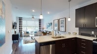 Located at 11501 W 81st St in Lenexa, Kansas, this single-floor residence showcases a thoughtfully designed open-concept layout that seamlessly connects the kitchen, dining, and living areas. The kitchen is a standout feature, outfitted with rich dark espresso cabinetry, a textured tile backsplash, stainless steel appliances including a range, microwave, and side-by-side refrigerator, and a generous island with a quartz countertop and undermount sink. Elegant pendant lighting illuminates both the island and the surrounding space. The living area is warm and inviting, highlighted by hardwood-style flooring, large windows with roller shades and patterned curtains that flood the room with natural light, and a ceiling fan overhead. The bedroom offers a serene retreat with plush carpeting, expansive windows overlooking a private balcony, and ample natural light. The bathroom features a dark-cabinetry vanity with a quartz countertop and well-appointed mirrors. Throughout the residence, crisp white walls provide a clean backdrop that complements the modern, cohesive design aesthetic, making this a refined and comfortable living space.