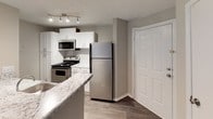 This single-story unit offers a thoughtfully designed open-concept layout that connects the living area and kitchen in a cohesive, functional arrangement. The living room is a standout space, featuring vaulted ceilings that lend a sense of openness and volume. A ceiling fan is centrally mounted, adding both comfort and style. Soft carpet flooring runs throughout the living and bedroom areas, while the kitchen transitions to wood-look hard flooring. A sliding glass door with vertical blinds opens to a private balcony, allowing natural light to fill the living space.

The kitchen is well-appointed with white shaker-style cabinetry, granite-inspired countertops extending to a breakfast bar peninsula, and stainless steel appliances including a refrigerator, gas range, and built-in microwave. Track lighting overhead provides focused illumination. A closet near the kitchen entry offers convenient storage.

The bedroom is a generously sized, carpeted retreat with a window that brings in natural light. The bathroom features a marble-pattern countertop vanity, an undermount sink, and a large mirror, complemented by neutral-toned walls throughout. The overall ambiance is clean, modern, and move-in ready.