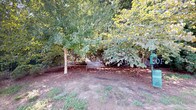 This single-floor space offers a thoughtful blend of comfortable interior living and dedicated outdoor amenity. The dog park is a standout feature, enveloped by mature, leafy trees that provide generous shade throughout the day. A classic metal bench is nestled among the trees on a mulched ground surface, creating a tranquil retreat for pet owners. A perimeter fence ensures a secure environment for pets to roam freely.

Inside, the interior rooms are finished with warm, neutral tones and plush carpeting, lending a clean and inviting atmosphere. White panel doors and crisp white baseboards provide a polished contrast against the soft beige walls. One room features a window with horizontal blinds that frames a pleasant outdoor view of lush greenery, allowing natural light to fill the space. The hallway connects seamlessly to an open-concept kitchen area, highlighted by dark cabinetry, a granite-topped island with decorative column details, and elegant pendant lighting overhead. Hardwood-style flooring in the common areas adds warmth and continuity throughout the layout. Together, the interior and outdoor elements create a well-rounded living environment suited for residents and their pets alike.