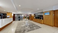Located at 1212 Avenue des Pins Ouest in Montréal, this sophisticated building lobby presents a polished and welcoming first impression. The space is organized into two seamlessly connected zones: a formal reception area and a comfortable lounge, all unified by a refined contemporary design. 

The reception area anchors the space with a sleek white marble-fronted desk backed by warm wood paneling, complemented by a built-in credenza with large mirror panels and integrated display screens. A striking glass-top accent table set atop a textured area rug serves as a central decorative focal point, adorned with fresh floral arrangements that add a refined touch. The flooring throughout this zone features large-format polished tiles that reflect the recessed lighting above.

A distinctive oval tray ceiling detail adds architectural interest overhead, drawing the eye upward and lending the space a sense of volume and grandeur. Transitioning into the lounge area, the flooring shifts to a neutral-toned carpet, where a dark sectional sofa with accent pillows and a low-profile coffee table create an inviting seating arrangement. Sheer floor-to-ceiling curtains filter natural light from the glazed entrance vestibule, which features sleek black-framed glass doors. Dark marble wall panels near the elevator bank add a luxurious contrast, completing the lobby's polished and professional ambiance.