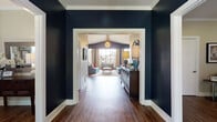 This thoughtfully designed single-floor space makes a strong first impression through a dramatic entryway corridor, where deep navy walls and crisp white trim frame a view into the open living area beyond. Rich hardwood flooring flows continuously throughout, anchoring the space with warmth and visual cohesion.

The expansive living area is defined by soaring vaulted ceilings and floor-to-ceiling windows that flood the room with natural light while offering outdoor views. A contemporary globe chandelier and a classic fireplace serve as focal points, complemented by layered seating arrangements and patterned area rugs that add texture and comfort. The bold navy accent walls provide striking contrast against the lighter ceiling and neutral tones throughout.

Adjacent to the living area, the dining space is equally well-appointed, featuring plantation shutters that allow for customizable light control, French doors opening to an outdoor balcony, and room for generous seating at a substantial dining table. Vibrant artwork adds a curated, welcoming character to the room.

The kitchenette is a standout feature, showcasing soft blue-grey cabinetry with brushed nickel hardware, white shiplap walls, light countertops, stainless steel appliances including a built-in microwave and refrigerator, and a striking large-scale painting as a focal centerpiece. Together, these spaces create a polished, inviting environment ideal for both gathering and everyday use.
